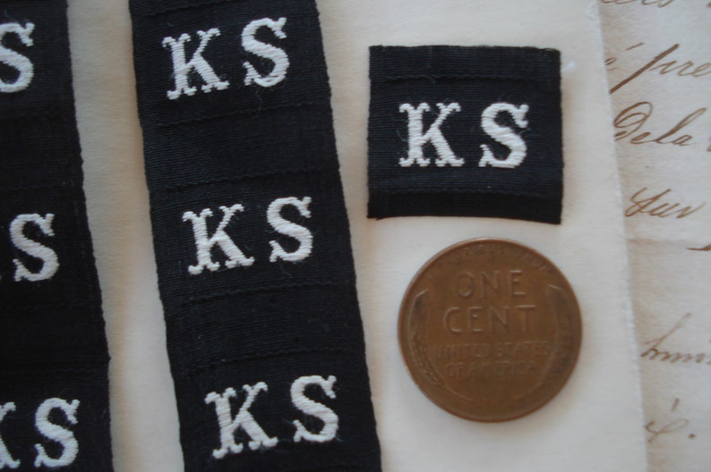 May include: Close-up of five black fabric labels with white embroidered letters spelling "KS". A single copper penny is also visible in the image.