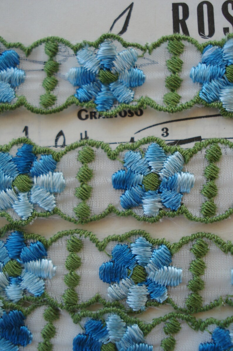May include: White lace trim with blue and green embroidered flowers. The trim has a scalloped edge.