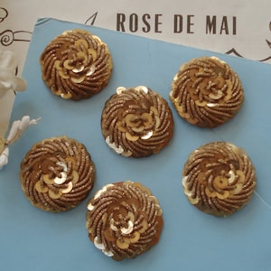 Può includere: Sei abbellimenti rotondi in tessuto color oro con un design a spirale. Ogni abbellimento è fatto di paillettes e filo.