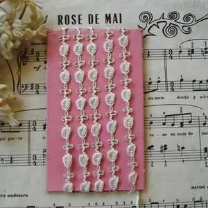 May include: White crocheted trim with a delicate floral design. The trim is displayed on a pink background.