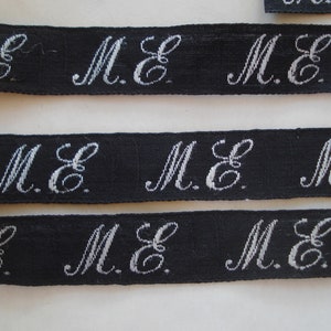 May include: Black ribbon with white embroidered letters that read "M.E." repeated across the ribbon.