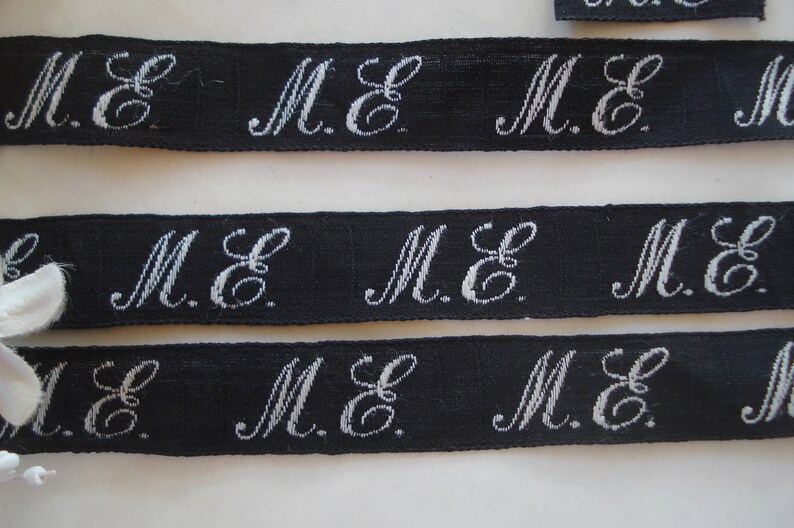 May include: Black ribbon with white embroidered letters that read "M.E." repeated across the ribbon.