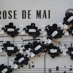 May include: Black and white trim with a floral design. The trim is made of fabric and has a ruffled edge. The trim is on a piece of sheet music with the words "ROSE DE MAI" printed on it.