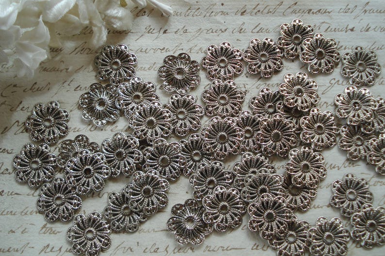 May include: A collection of silver metal filigree embellishments in the shape of flowers. The embellishments are delicate and intricate, with a floral design.