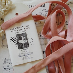 Könnte beinhalten: Eine Rolle mit 9,14 Metern staubigem rosa Satin-Rückseiten-Samt mit gewebtem, schnell laufendem Rand. Das Band ist aus 100% Rayon und wurde in der Schweiz hergestellt. Das Etikett zeigt auch eine Schwarzweißillustration einer Ballerina.