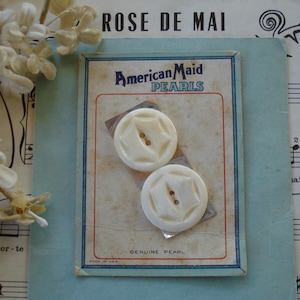May include: Two off-white, star-shaped buttons on a vintage card labeled "American Maid PEARLS." The card has a red and blue border and the text "GENUINE PEARL" and "MADE IN USA." The background includes sheet music and dried flowers.