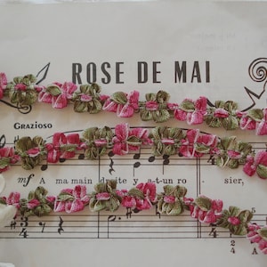 May include: A close-up of a pink and green fabric trim with a ruffled design. The trim is laid out on a sheet of music with the words "ROSE DE MAI" printed at the top.