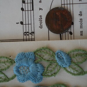 May include: A close-up of blue and green lace trim with floral designs. The lace is delicate and intricate, with small flowers and leaves.