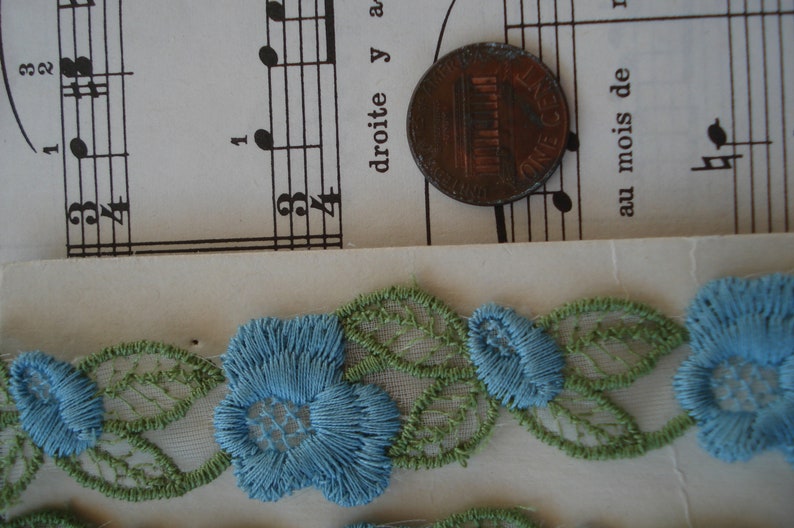 May include: A close-up of blue and green lace trim with floral designs. The lace is delicate and intricate, with small flowers and leaves.