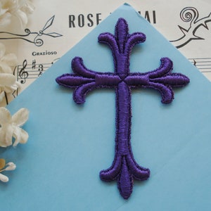 May include: A purple embroidered cross with ornate detailing, set against a light blue background. The cross is the focal point, showcasing a detailed stitch pattern. The background includes sheet music and floral accents.
