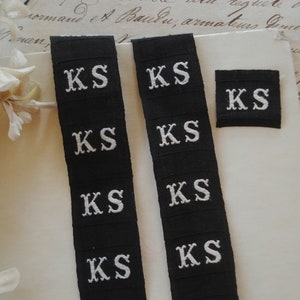 May include: Four black fabric labels with white embroidered letters spelling "KS" on each label. The labels are arranged in two rows of two, with the top row slightly offset from the bottom row.