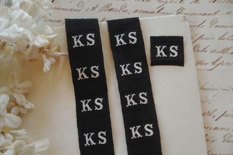 May include: Four black fabric labels with white embroidered letters spelling "KS" on each label. The labels are arranged in two rows of two, with the top row slightly offset from the bottom row.
