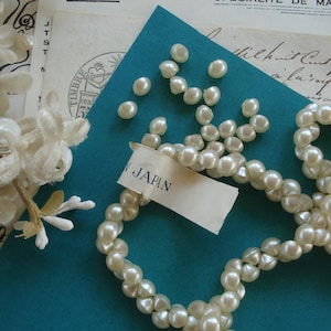 Peut inclure: Un collier de perles artificielles blanches sur un fond turquoise. Les perles sont disposées en cercle lâche avec une petite étiquette indiquant "Japon".