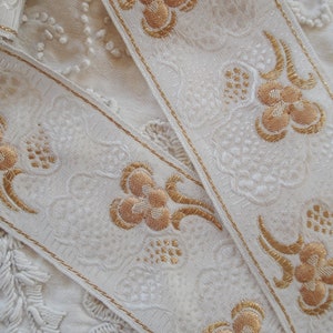 Pode incluir: Close-up de fita cor marfim com bordado floral dourado. A fita apresenta um padrão repetido de flores e folhas estilizadas, com acabamento dourado nas bordas. O fundo é um tecido texturizado branco sujo.
