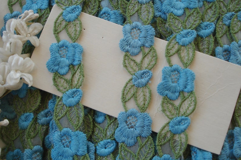 May include: Blue and green floral lace trim with a delicate floral pattern. The trim features small blue flowers with green leaves.