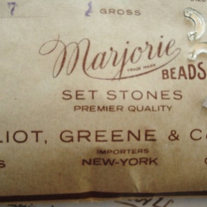 May include: A vintage label for Marjorie Beads, a company that sells set stones. The label is brown with white text and reads "Marjorie Beads, Set Stones, Premier Quality, Elliot, Greene & Co. Inc., Importers, New York, Gablon, Paris".
