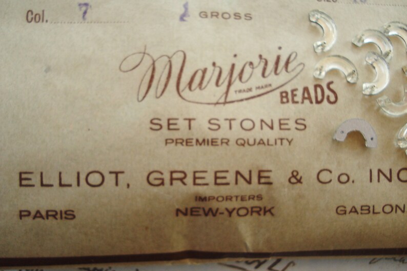 May include: A vintage label for Marjorie Beads, a company that sells set stones. The label is brown with white text and reads "Marjorie Beads, Set Stones, Premier Quality, Elliot, Greene & Co. Inc., Importers, New York, Gablon, Paris".