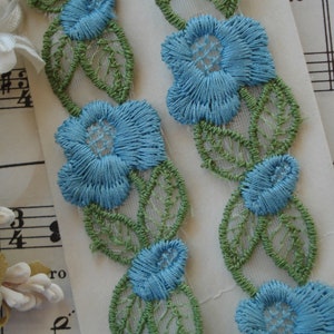 May include: Two pieces of blue and green floral lace trim with a white background. The lace trim features delicate blue flowers with green leaves.