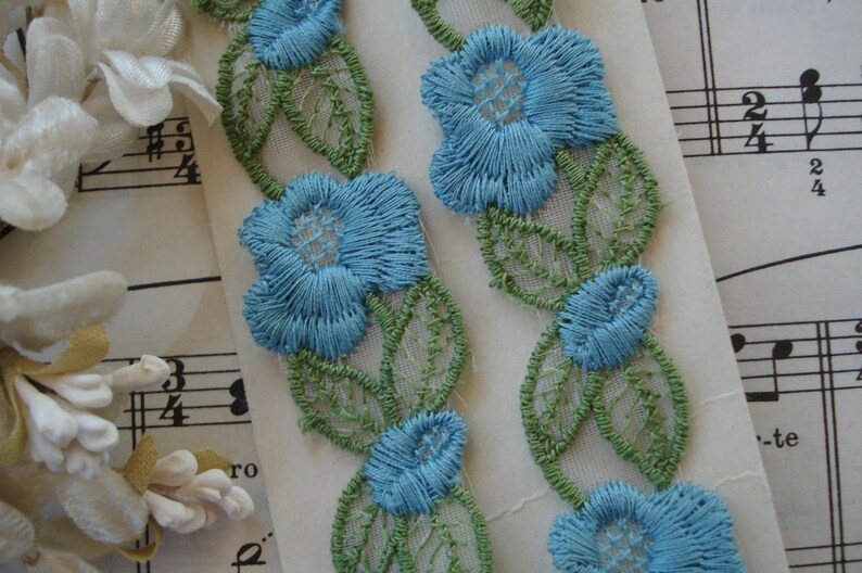 May include: Two pieces of blue and green floral lace trim with a white background. The lace trim features delicate blue flowers with green leaves.