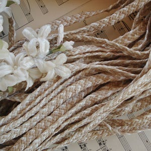 May include: Close-up of braided rope in shades of beige and cream, with a cluster of white fabric flowers. The rope is tightly woven, creating a textured surface. The flowers have delicate petals and are clustered together. The background features sheet music.