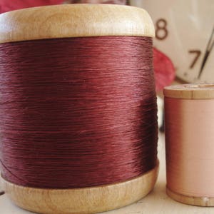 May include: Two spools of thread, one is a deep burgundy red and the other is a pale pink. Both spools are made of wood and have a vintage look.