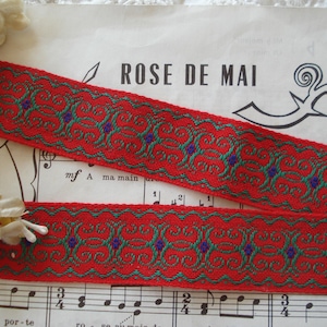 Könnte beinhalten: Roter und grüner gemusterter Besatz mit einem sich wiederholenden Muster. Der Besatz hat einen roten Hintergrund mit einem grünen und violetten Blumenmuster. Der Besatz befindet sich auf einem Blatt Musik mit dem aufgedruckten Text "ROSE DE MAI".