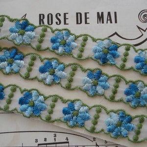 May include: Three rows of blue and white embroidered floral trim with green accents. The trim is on a white background and has a scalloped edge.
