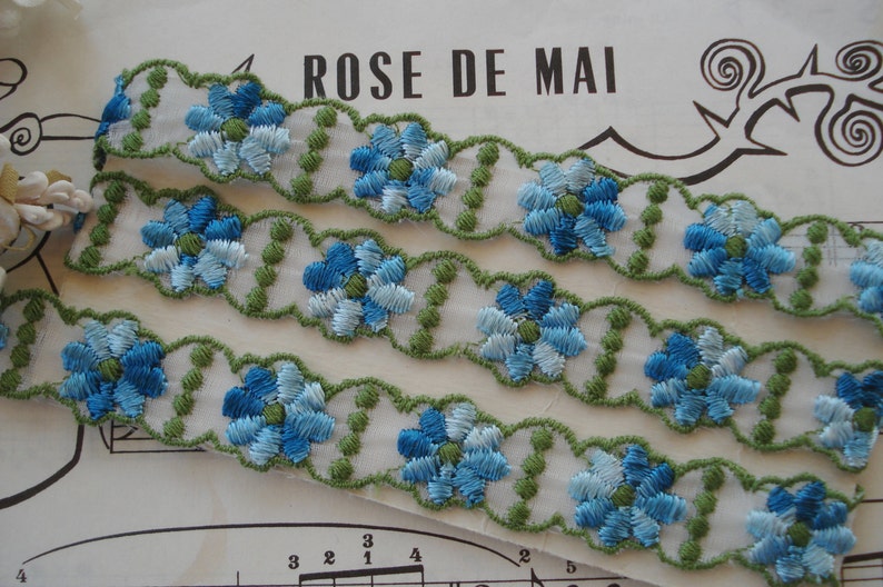 May include: Three rows of blue and white embroidered floral trim with green accents. The trim is on a white background and has a scalloped edge.