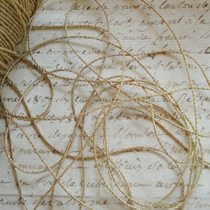 Op de afbeelding: Een close-up van een gouden metalen koord of trim, verward en overlappend op een stuk papier met handgeschreven tekst. Het koord is dun en heeft een subtiele glans.