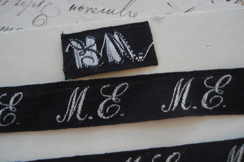 May include: A black fabric label with white embroidered letters "M.E." and a small black fabric label with a white embroidered image of two shoes.