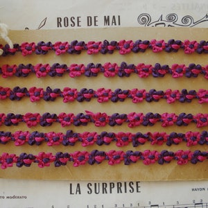 May include: Five rows of pink and purple fabric trim with a flower design. The trim is displayed on a brown card with the text "ROSE DE MAI" and "LA SURPRISE" printed on it.