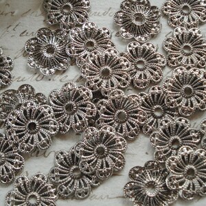 May include: A collection of silver filigree metal embellishments with a floral design. The embellishments are arranged in a random pattern on a white surface.