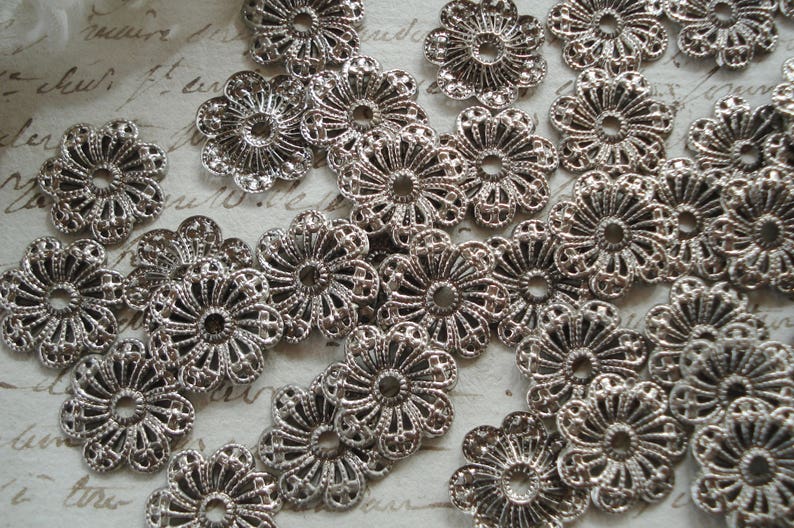 May include: A collection of silver filigree metal embellishments with a floral design. The embellishments are arranged in a random pattern on a white surface.