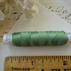 May include: A spool of green thread, labelled "Ideal Soie" and "Gütermann", lies on a white surface. A wooden ruler with the word "THREADS" is in the foreground. The thread is approximately 4 cm long and is a muted green colour.