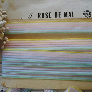 May include: Pastel striped ribbon in shades of pink, blue, yellow, and green. The ribbon is layered on a piece of aged paper with the text "ROSE DE MAI" visible. Dried flowers and sheet music are in the background.