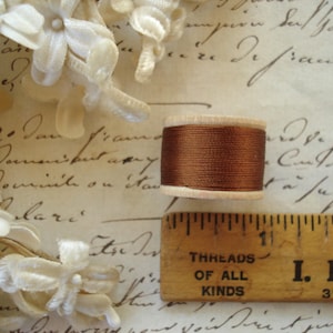 May include: A spool of dark brown thread with a light-colored wooden core, positioned next to a wooden ruler that shows the spool is approximately 1 inch in diameter. The thread is tightly wound around the center of the wooden spool.