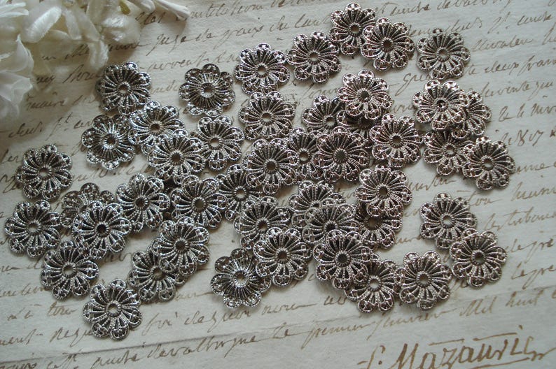May include: A large quantity of silver metal flower-shaped embellishments with intricate designs. The embellishments are scattered on a white surface with handwritten text.
