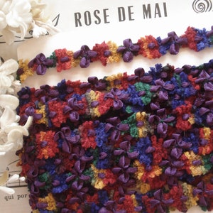 May include: Multicolored fabric trim with small flower shapes in shades of red, purple, blue, green, and yellow. The trim is made of a fuzzy material and has a slightly raised texture. The text "ROSE DE MAI" is visible in the background.