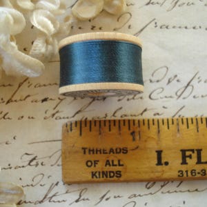 May include: A wooden spool of dark teal thread positioned above a wooden ruler. The ruler shows the spool has a diameter of approximately 1 inch. The spool features light wood edges and a dark teal thread wrapped around the center cylinder.