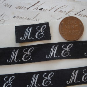 May include: Close-up of several black and white fabric ribbon trims with the initials "M.E." embroidered in white. The ribbon trims are arranged in a staggered pattern on a white surface.