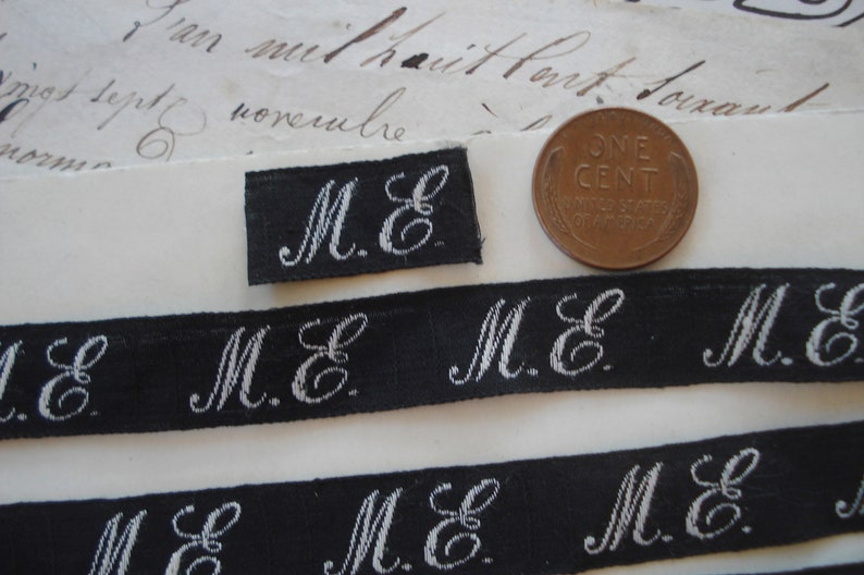 May include: Close-up of several black and white fabric ribbon trims with the initials "M.E." embroidered in white. The ribbon trims are arranged in a staggered pattern on a white surface.
