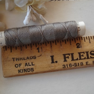 May include: A spool of grey thread with a white label, next to a vintage wooden ruler. The ruler is marked with inches and has text that reads "THREADS OF ALL KINDS" and "I. FLEISCHE". The thread is approximately 2.5 cm in length.