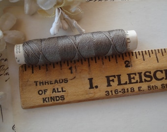 May include: A spool of gray thread with a white label, next to a vintage wooden ruler. The ruler is marked with inches and has text that reads "THREADS OF ALL KINDS" and "I. FLEISCHE". The thread is approximately 1 inch in length.