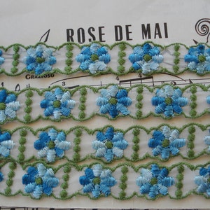 May include: Three rows of blue and green embroidered floral trim. The trim is on a white background and has a scalloped edge.