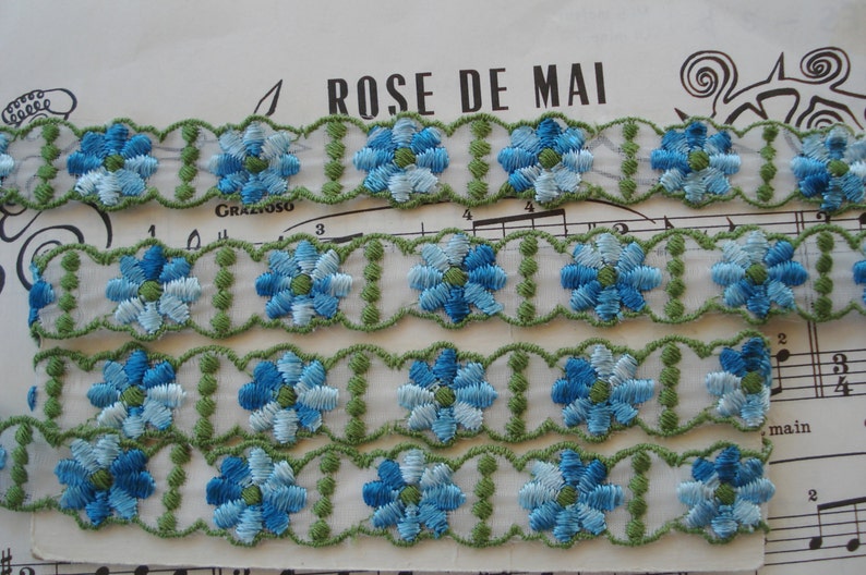 May include: Three rows of blue and green embroidered floral trim. The trim is on a white background and has a scalloped edge.