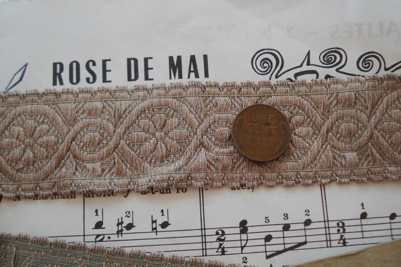 Puede incluir: Un primer plano de un ribete de tela con estampado marr&oacute;n y beige con una moneda de un centavo encima. La tela tiene un dise&ntilde;o tejido y un borde festoneado. La moneda es de color cobre y tiene las palabras "ONE CENT" inscritas en ella.
