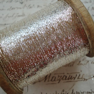 May include: A spool of silver metallic thread wound around a wooden bobbin. The thread has a textured, woven appearance.
