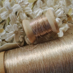 May include: Two spools of gold metallic thread, one on a wooden spool and one on a larger spool. The spools are surrounded by white fabric flowers.