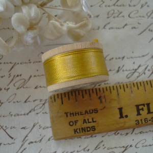 May include: A wooden spool wrapped with metallic gold thread, positioned next to a yellow wooden ruler that shows the spool has a diameter of approximately 2.5 cm. The spool is light-coloured wood with a smooth, cylindrical shape.