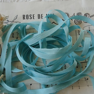 May include: A skein of light blue ribbon on a sheet of music with the words "ROSE DE" visible. The ribbon is a soft, silky material and is perfect for crafting or decorating.